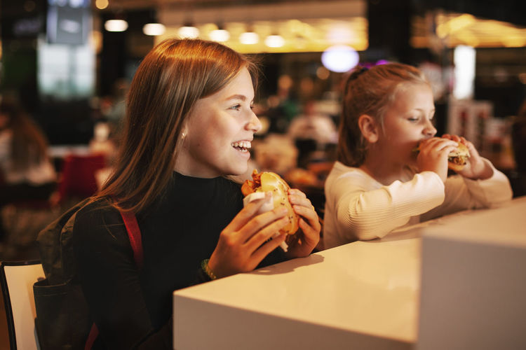 Beautiful schoolgirl eating hamburger sitting in a cafe indoors. happy child eating junk food