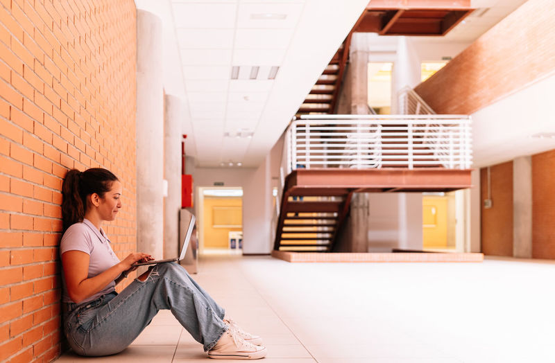 Full body side view of delighted female student in casual wear surfing internet on netbook while sitting on floor near brick wall