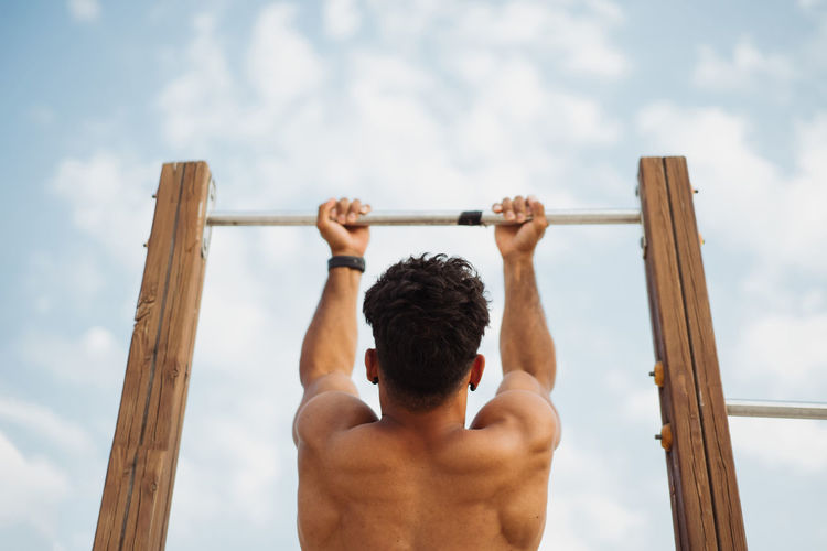 Low angle back view of unrecognizable muscular hispanic ethnic male athlete with naked torso doing chin ups on horizontal bar during training on background of clear blue sky