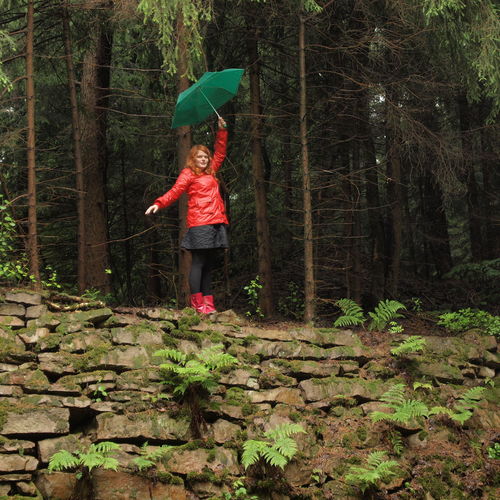 Young woman in raincoat with umbrella standing on stone wall at forest