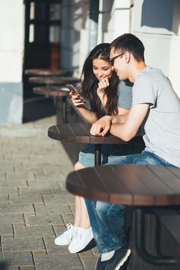 Young woman with man using phone while sitting at sidewalk cafe on sunny day