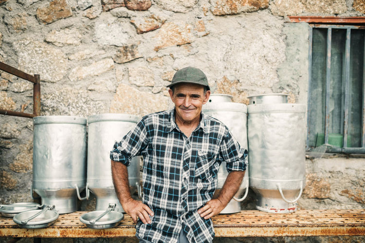 Smiling male goat herder standing with hands on hip in front of milk canisters at farm