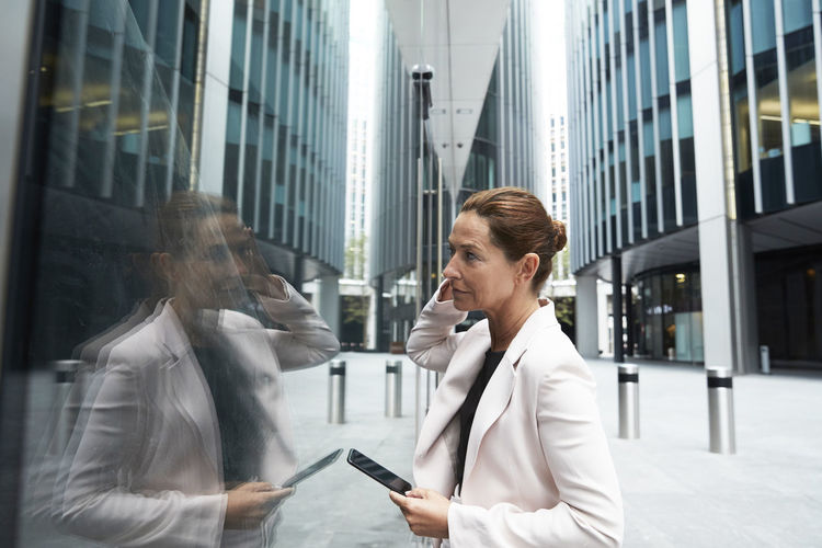 Mature female entrepreneur holding smart phone while looking at her reflection on office building glass window