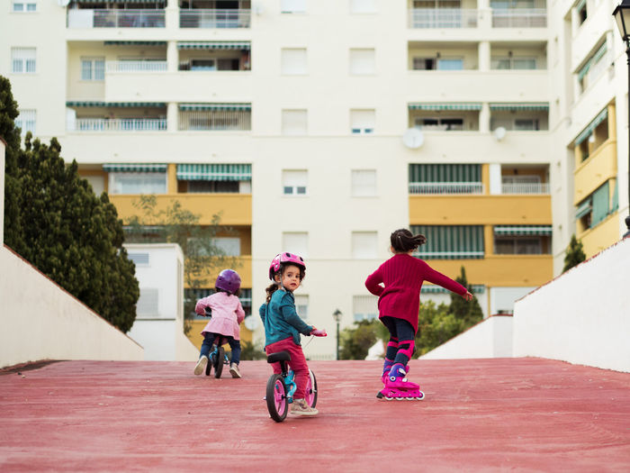 Little girl on bicycle looking at camera over shoulder while spending time with friends near modern apartment building