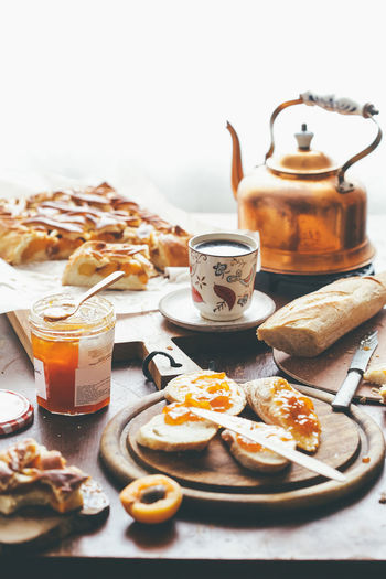 Cup and copper teapot of hot tea placed on table amidst pie and toasts with apricots and jam at home