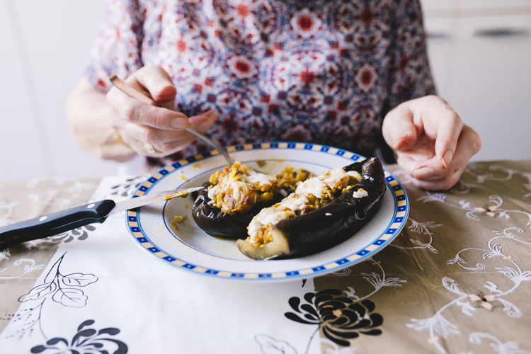 Senior woman eating stuffed eggplant in the kitchen, partial view