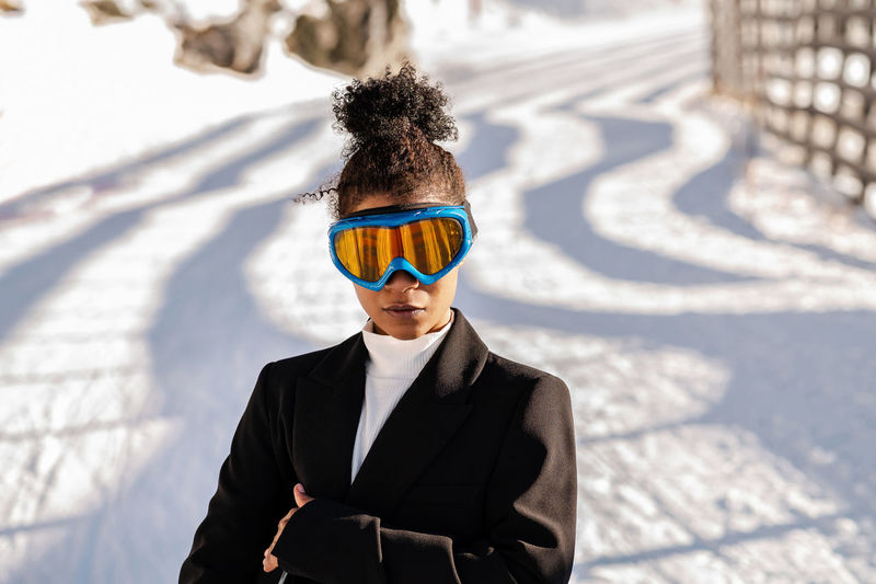 Stylish young ethnic female athlete in protective glasses looking at camera on snowy roadway in sunlight
