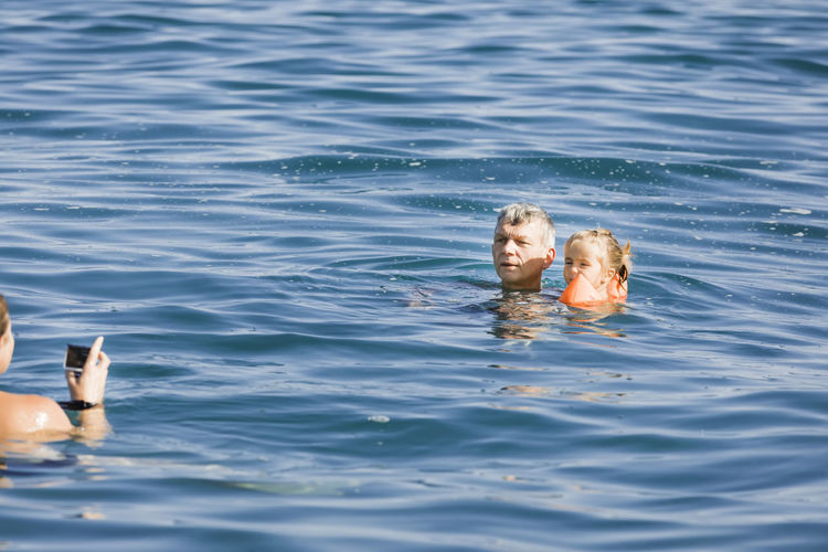 Woman photographing grandfather and granddaughter swimming in sea