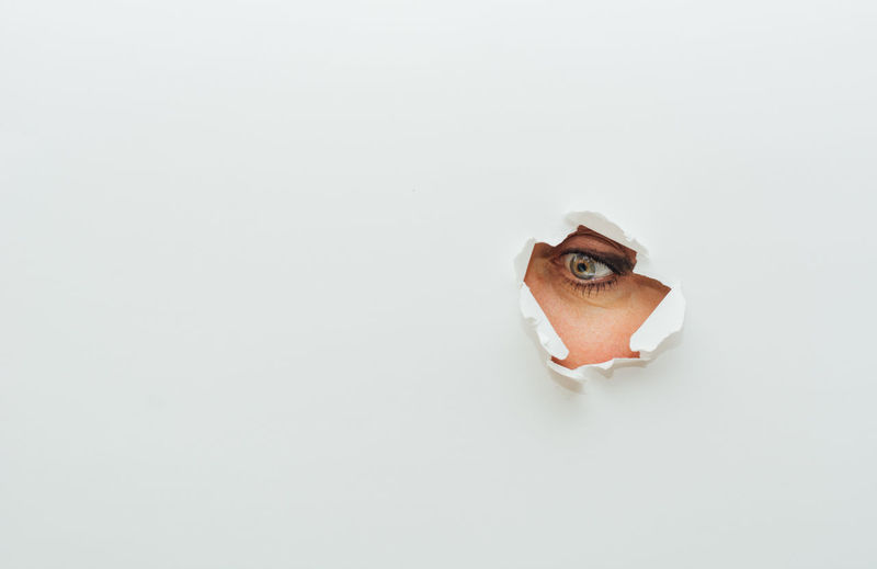 Blue eye of unrecognizable female looking away through ripped hole in white paper cardboard background person