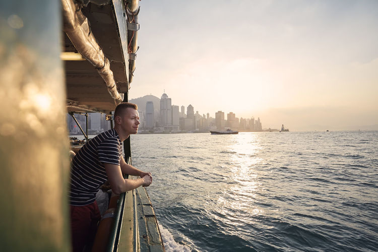 Pensive man looking from ferry boat against urban skyline st beautiful sunset. tourist in hong kong.