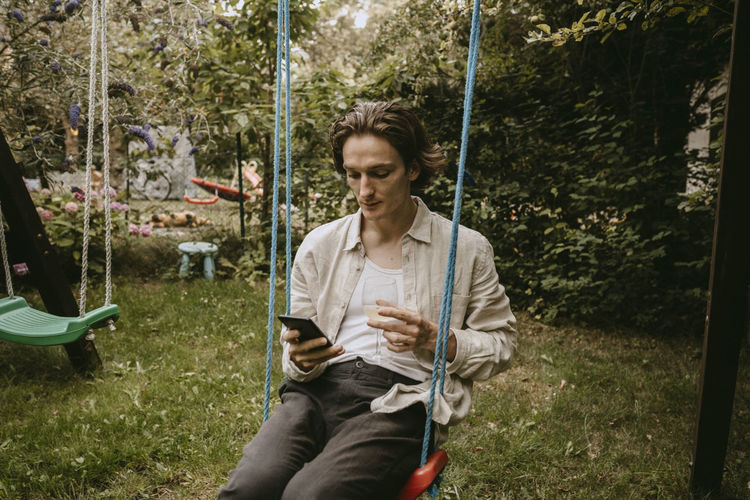Contemplating man with drink using smart phone while sitting on swing in yard
