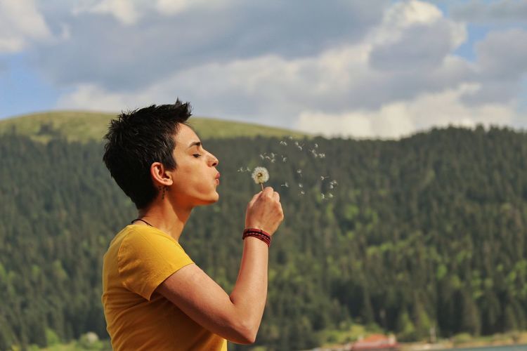 Side view of young woman blowing dandelion against tree mountain
