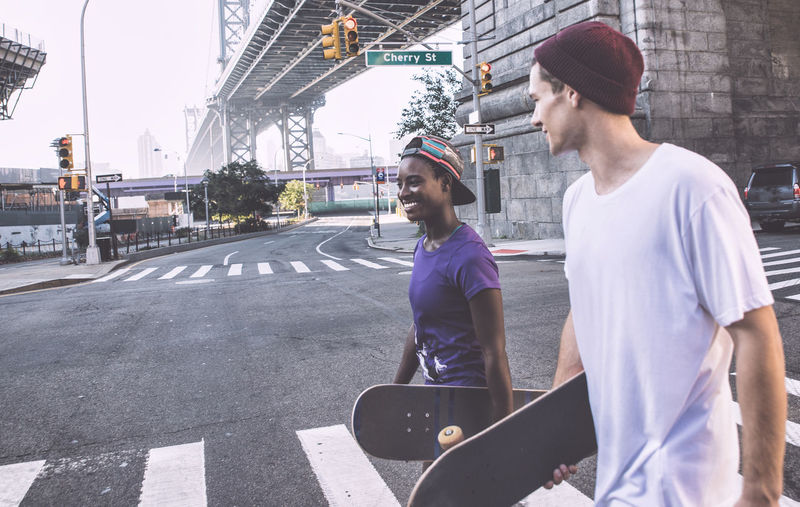 Young man with woman walking on road while holding skateboard