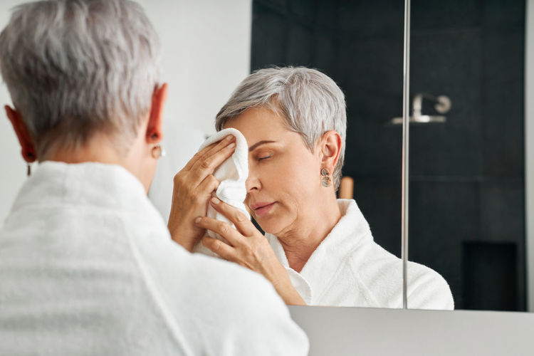 Senior woman wiping face while reflecting in mirror