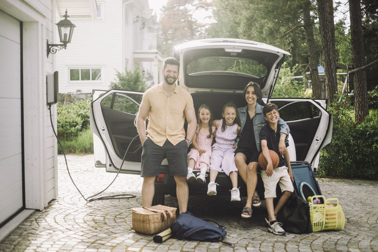 Portrait of happy family sitting in electric car trunk outside house