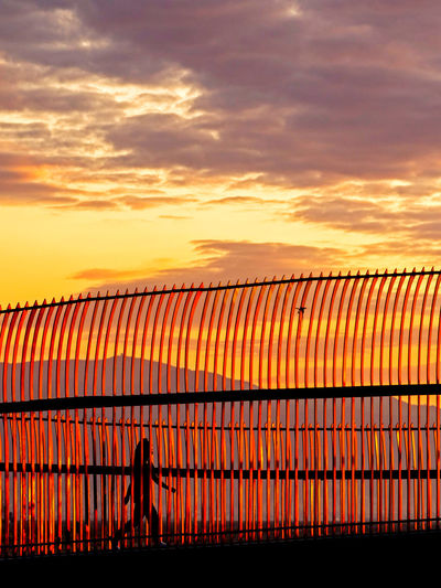 Silhouette person walking behind fence against orange sky