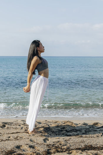 Side view of ethnic female leaning back while standing in ashta chandrasana pose during yoga practice on sandy coast against ocean