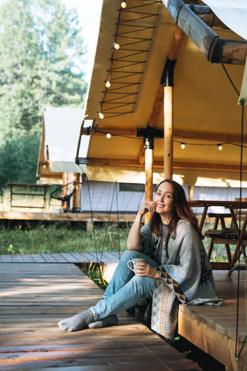 Young brunette woman in poncho drinking tea and relaxing in glamping in nature