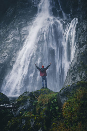 Rear view of man with arms raised standing on rock by waterfall