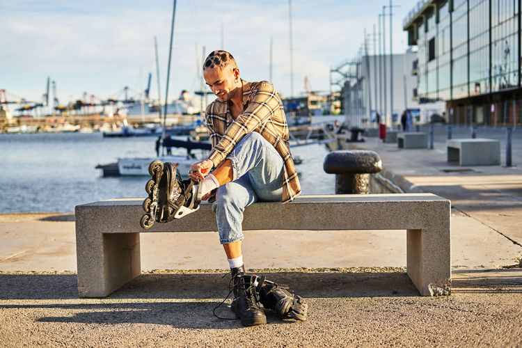 Full body of eccentric male sitting on bench and putting on roller skates on waterfront for riding at spare time