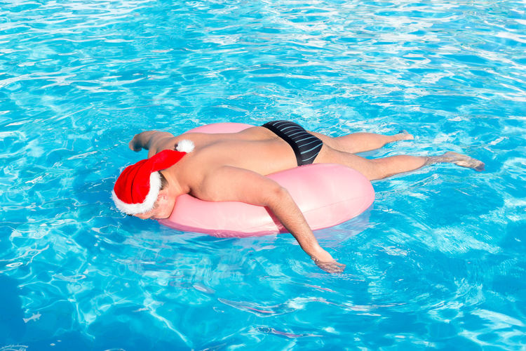 Shirtless man wearing santa hat while lying on inflatable ring in pool