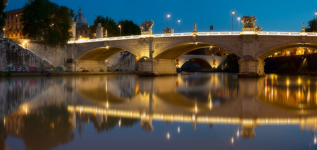 Reflection of bridge on river against sky in city at night