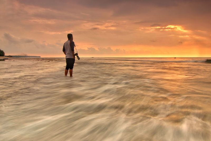 Rear view of man standing at beach against cloudy sky during sunset