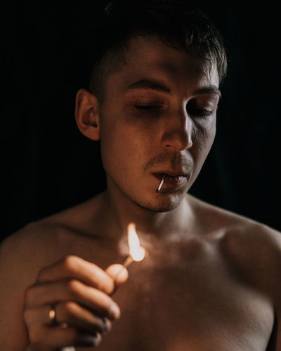 Close-up of shirtless man holding lit matchstick against black background
