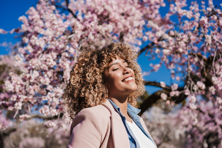Smiling hispanic woman with eyes closed in park enjoying sunny day. spring flowers background
