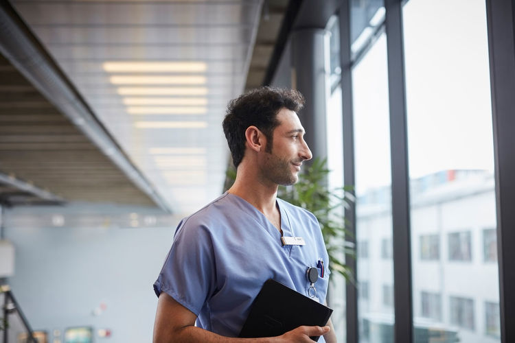 Thoughtful young male nurse holding digital tablet while looking through window at hospital