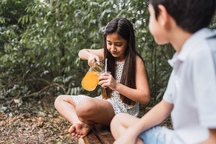 Cheerful child pouring lemonade into calabash gourd while sitting with crossed legs against content sibling on bench in afternoon