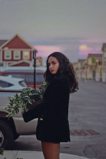 Side view portrait of teenage girl holding plants standing on street during sunset