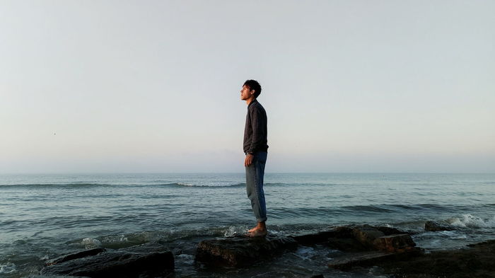 Side view of man standing on rock in sea against clear sky