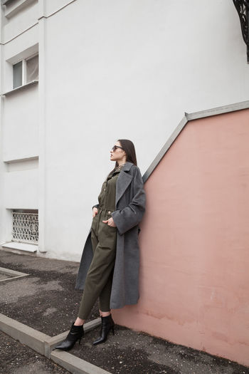 Full length side view portrait of young woman in grey coat and suit against wall outdoors, outwear 