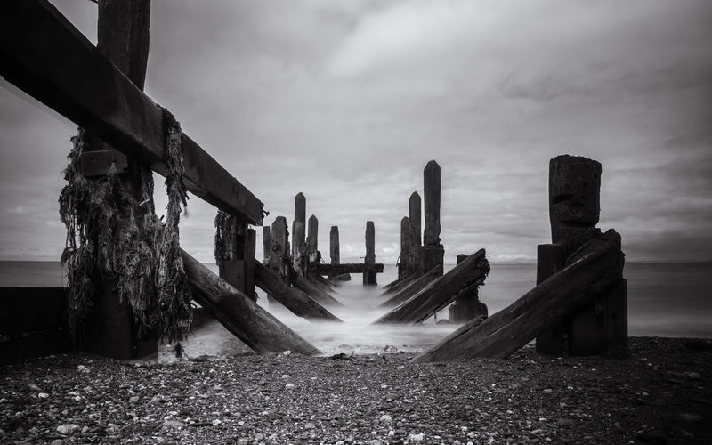 Abandoned wooden structure at beach against cloudy sky