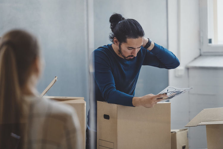 Young businessman reading book while unpacking cardboard box in new office