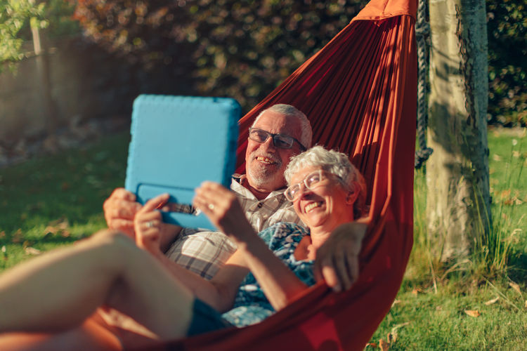 Smiling senior couple lying on hammock outdoors in backyard using tablet