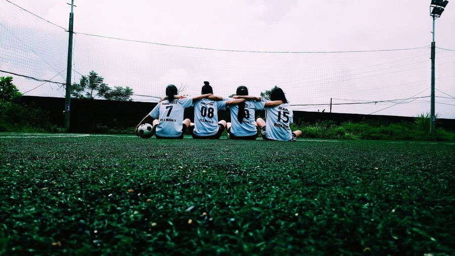 Rear view of soccer players sitting on field against sky