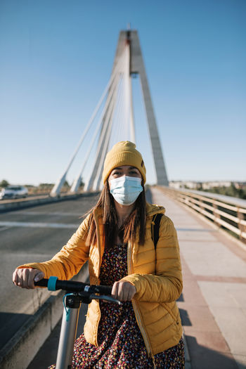 Woman in protective face mask with electric scooter on bridge in city during covid-19