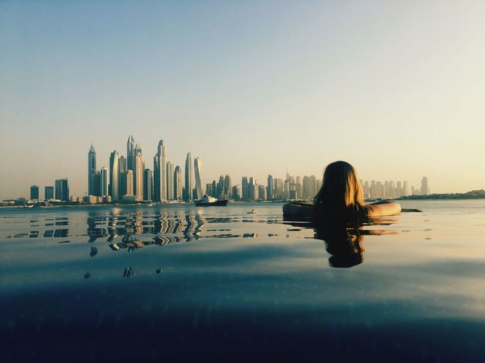 Rear view of woman looking at modern building while swimming in infinity pool against clear sky during sunset