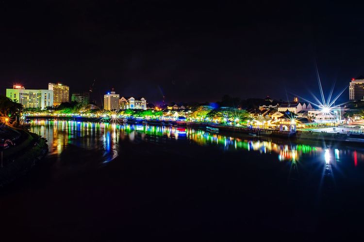 Illuminated buildings by river against sky at night