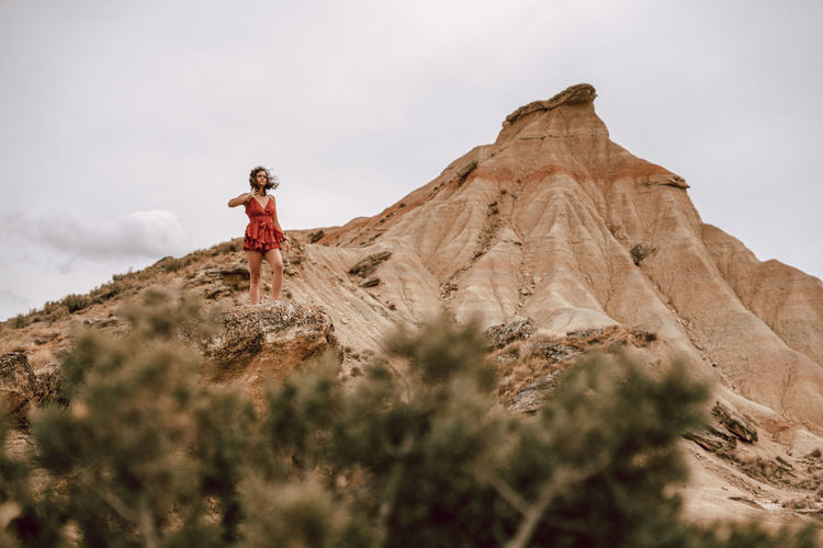 LOW ANGLE VIEW OF WOMAN STANDING ON ROCKS AGAINST MOUNTAIN