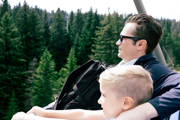 Happy little boy and his father riding on ski lift above the forest in summer day.