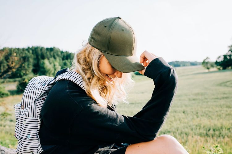 Woman sat in the countryside holding her cap over her face looking shy