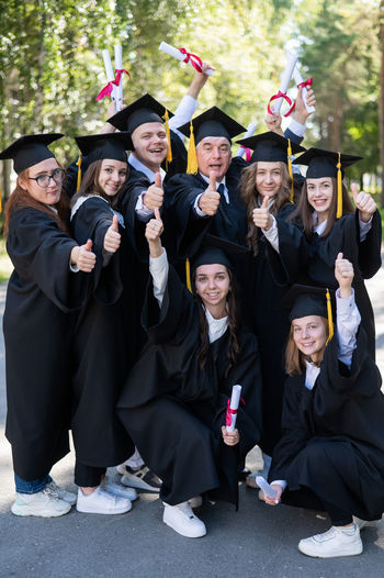 A group of graduates in robes give a thumbs up outdoors. elderly student. vertical
