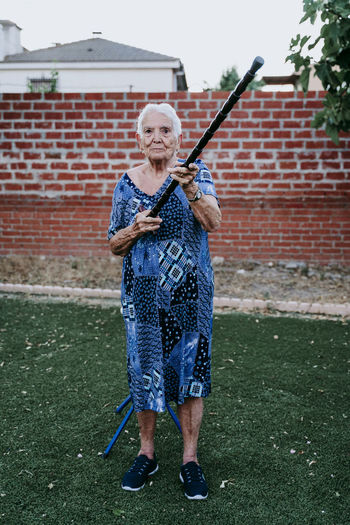 Full length of senior woman holding stick standing against brick wall