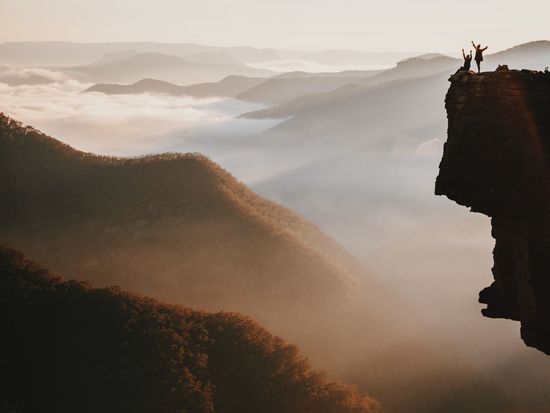 Silhouette people standing on cliff with arms raised by mountains against sky