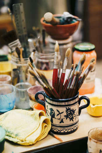 From above of table with cans with colors and paint brushes located around white ceramic cup with blue patterns and brushes inside