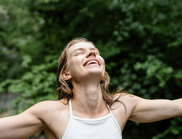 Woman turning her face to the sky and laughing, eyes closed. green trees on the background