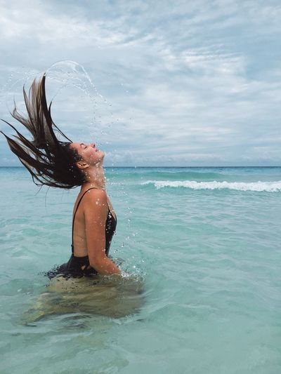 Side view of young woman tossing wet hair in sea against sky